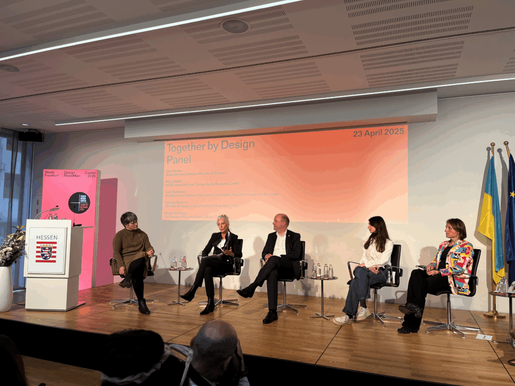 Redner einer Podiumsdiskussion für together by design auf der Bühne sitzend, mit der Präsentation hinter ihnen
